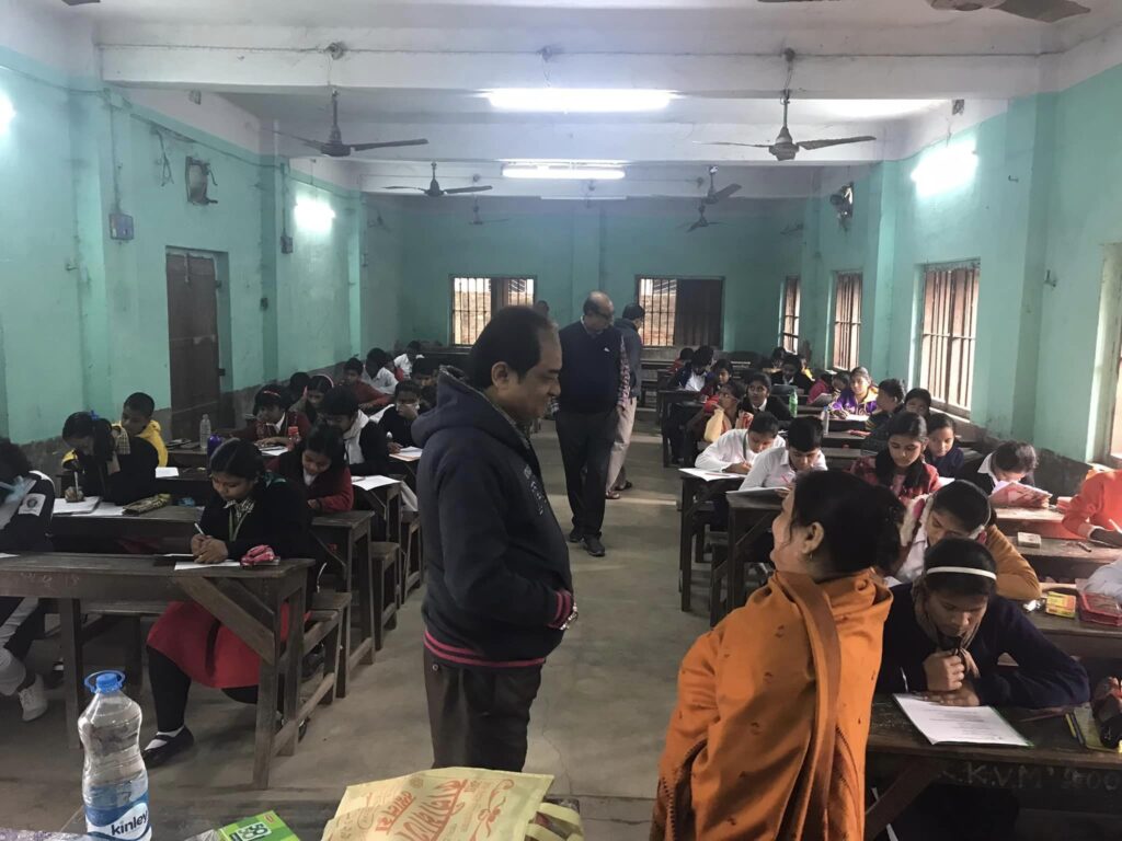 An invigilator stands between rows of wooden desks in a green-walled hall while students write their examination papers.