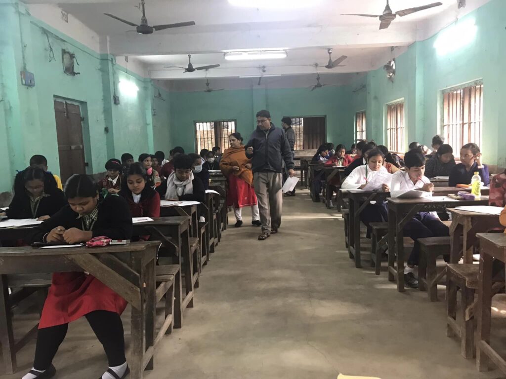 Students in school uniform write at wooden desks in a wide green-walled hall while two invigilators walk the aisle.