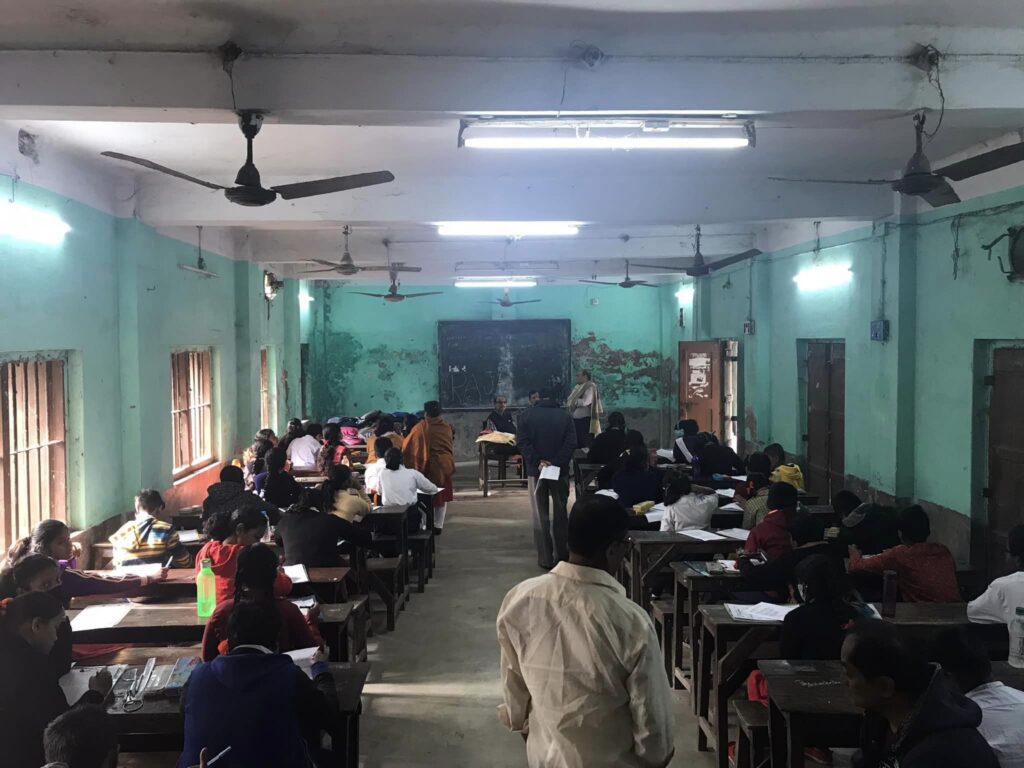 A long view down an examination hall, students at wooden desks on both sides of a central aisle and invigilators standing near the blackboard.