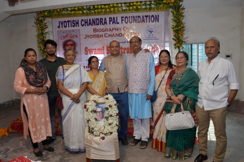 Family members and guests stand together behind a garlanded framed portrait of Jyotish Chandra Pal.