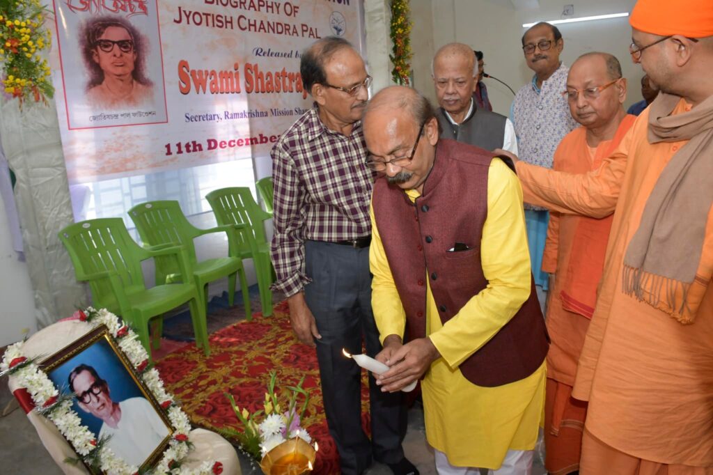 A guest bends to light a lamp beside the garlanded portrait of Jyotish Chandra Pal while monks and members look on.