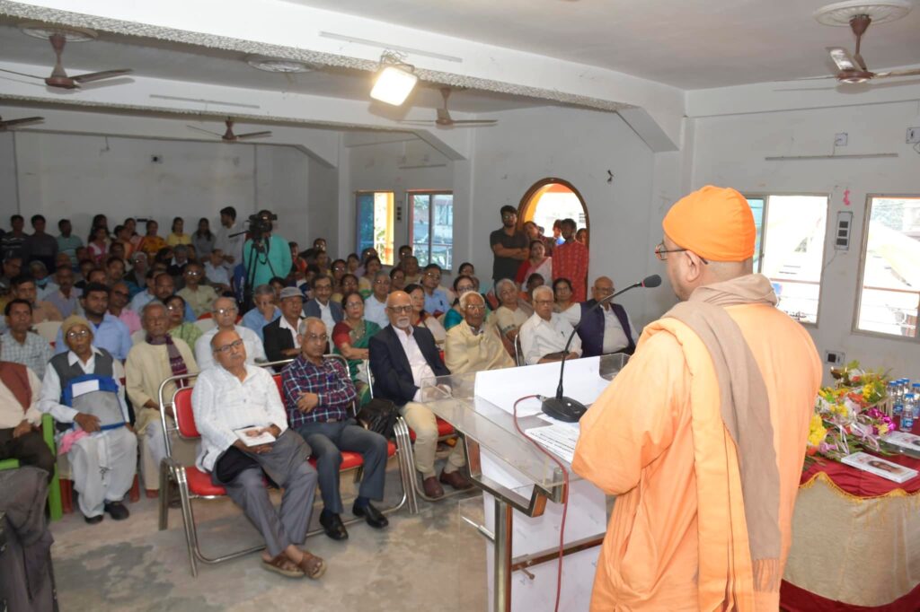 A monk in saffron robes addresses a seated audience of about sixty people from the podium.