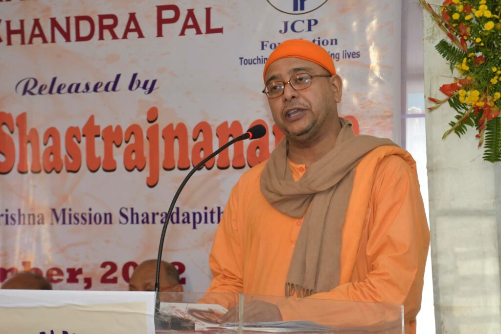 A monk in saffron robes and cap speaks from the podium beneath the banner announcing the release.