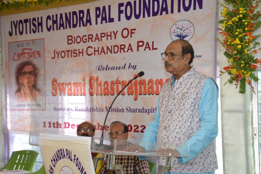 A speaker in a blue patterned waistcoat addresses the gathering from the podium, the banner behind him dated 11 December 2022.