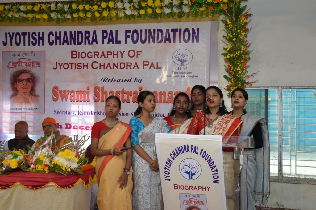 Six young women in saris sing together at the microphone in front of the launch banner, with seated guests to one side.