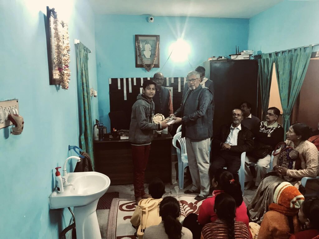 A boy in a grey hooded jacket accepts a trophy from an elderly guest as members seated along the wall look on.