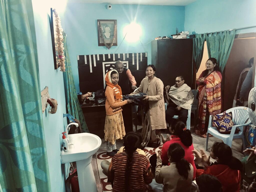 A girl in an orange hooded jumper accepts a backpack from a woman guest while the room applauds.
