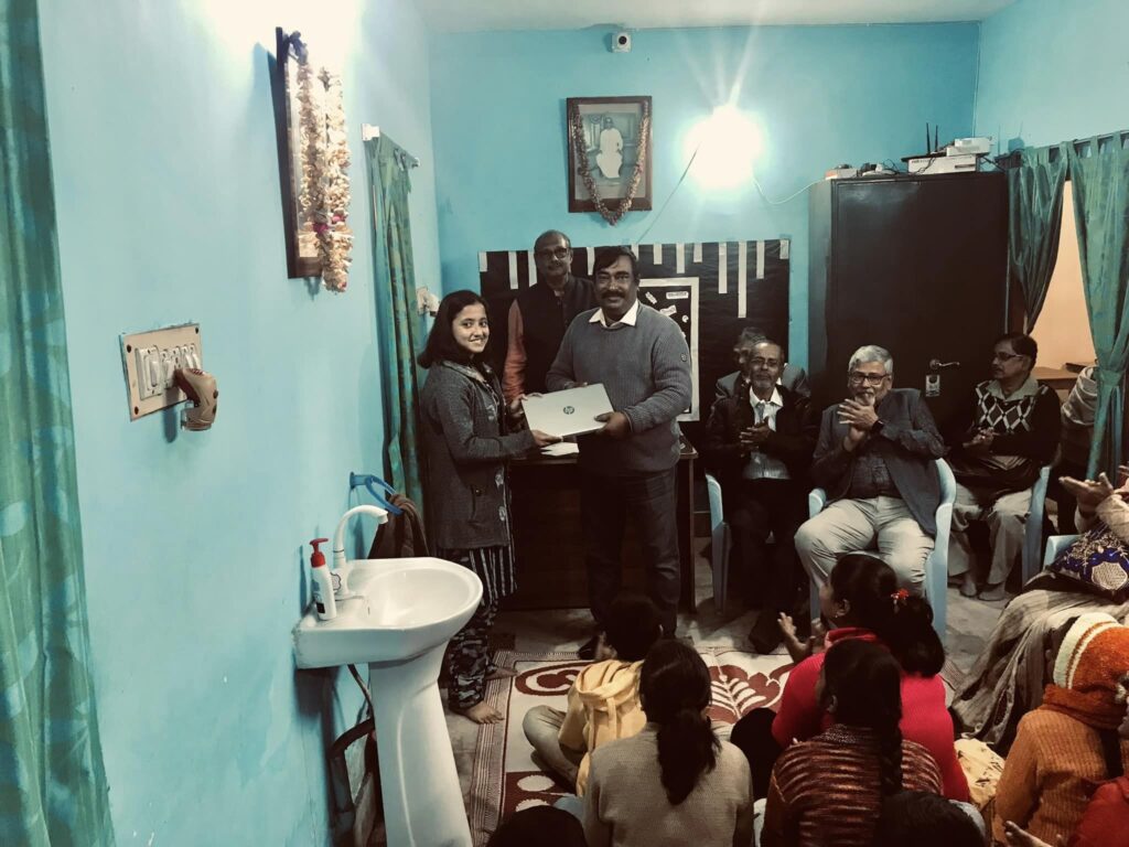 A student receives a laptop from a Foundation member at the prize ceremony, watched by seated guests and students sitting on the floor.