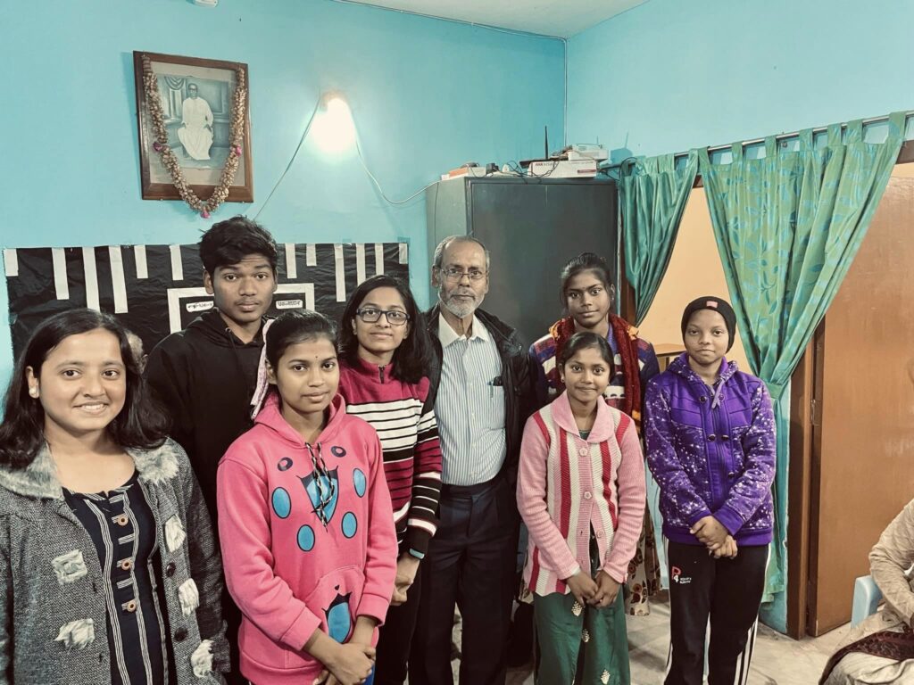 Seven students stand with an elderly Foundation member for a photograph beneath a garlanded portrait.