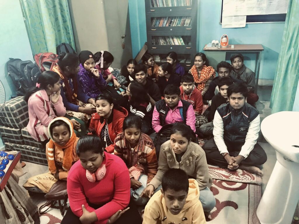 About twenty students sit closely together on the floor of a small room lined with a bookcase, waiting for the ceremony.